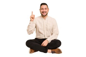 Adult latin man sitting on the floor cut out isolated showing number one with finger.