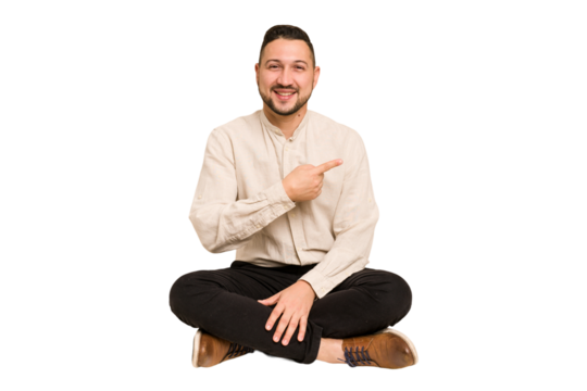 Adult latin man sitting on the floor cut out isolated smiling and pointing aside, showing something at blank space.