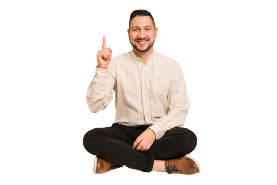Adult latin man sitting on the floor cut out isolated showing number one with finger.