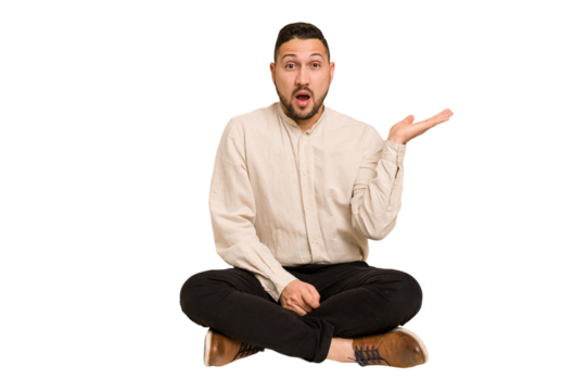 Adult latin man sitting on the floor cut out isolated impressed holding copy space on palm.