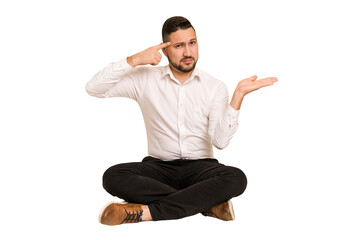 Adult latin man sitting on the floor cut out isolated holding and showing a product on hand.