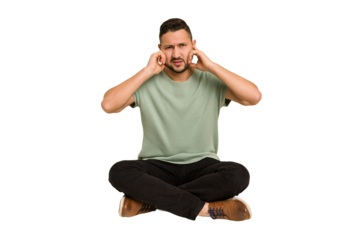 Adult latin man sitting on the floor cut out isolated covering ears with fingers, stressed and desperate by a loudly ambient.