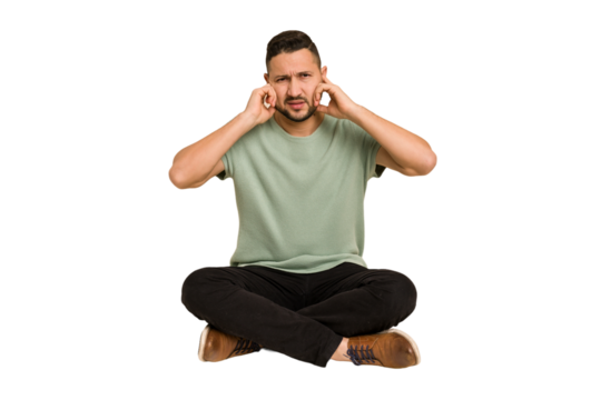Adult latin man sitting on the floor cut out isolated covering ears with fingers, stressed and desperate by a loudly ambient.