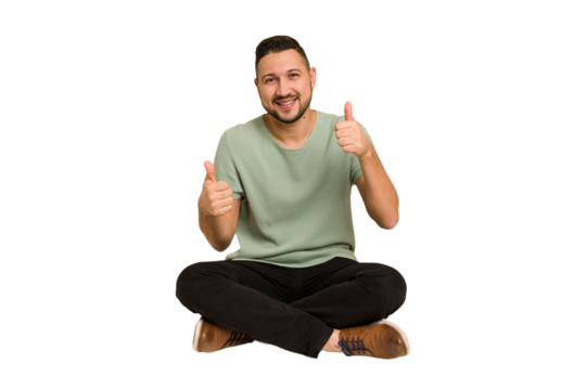 Adult latin man sitting on the floor cut out isolated raising both thumbs up, smiling and confident.