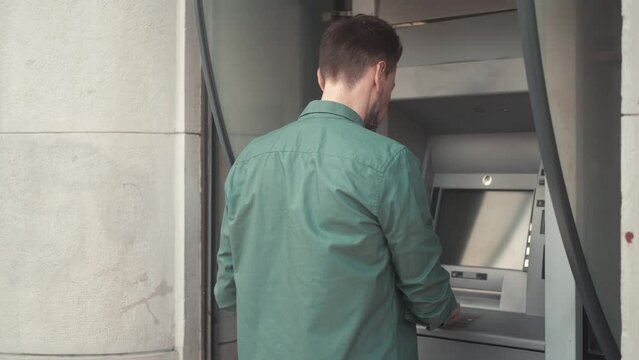 Back view portrait of young adult caucasian man using ATM terminal making financial operations on street. Beautiful concentrated male inserting bank card and entering pincode to withdraw money.