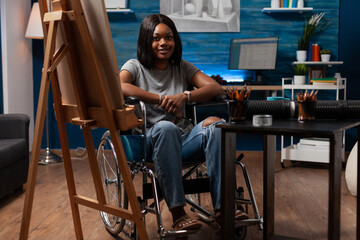 Young african american female professional artist wheelchair user looking at camera from home art studio. Portrait of woman cartoonist surrounded by creative tools in atelier at home.