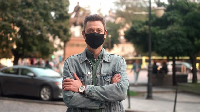 Portrait of confident serious young caucasian man standing with crossed arms in city center looking at camera wearing medical mask. Handsome adult male wearing casual clothes in urban beautiful park.