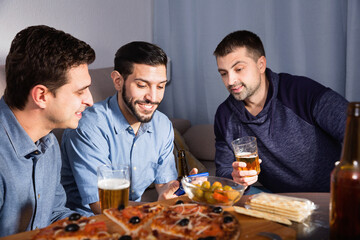 Three cheerful positive smiling men using phone while enjoying beer and pizza at home