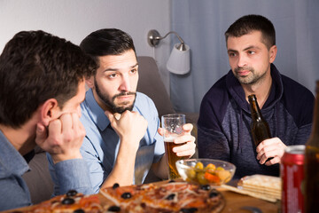 Three young unhappy men discussing problems while drinking beer with pizza at home