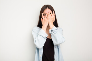 Young caucasian woman isolated on white background blink at the camera through fingers, embarrassed covering face.