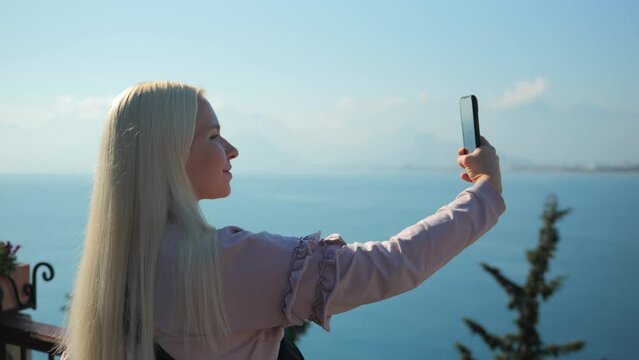 A young pretty blonde woman stands on the observation deck by the sea and takes pictures of landscapes on her phone. High quality 4k footage