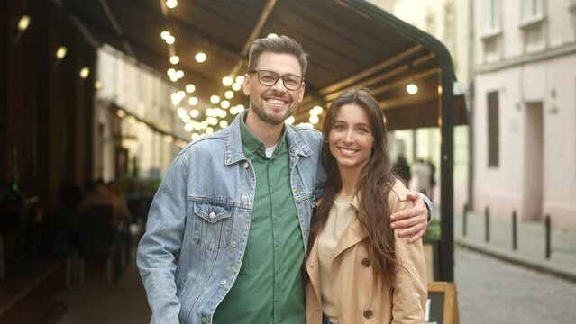 Portrait of lovely cute couple of two young people having date near cozy cafe in city center. Pretty beautiful woman standing near handsome cheerful man hugging looking at camera smiling.