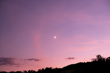 Lune dans un ciel rose, forêt reconnaissable en dessous. Nuages roses