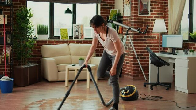 Young woman sweeping dirt with mop in living room, mopping wooden floors and cleaning apartment. Casual female person washing tiles with cleaner, being focused on housework or chores.