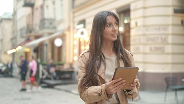 Portrait of cute young caucasian woman using tablet drawing online standing outside looking at beautiful city. Attractive pretty female designer using technologies standing in city center.