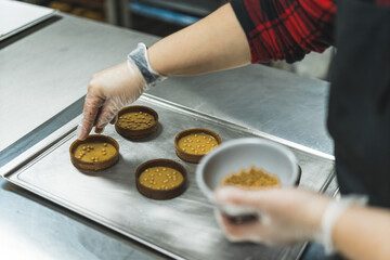 Top view of tarts with caramel icing being decorated with golden pearls on a baking tray by a expert wearing transparent gloves. Blurred foreground. High quality photo
