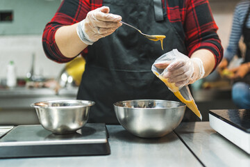 Front view of a expert putting caramel icing into a transparent pastry bag from a metal bowl with a spoon. Two metal bowls placed next to each other on a kitchen counter. High quality photo