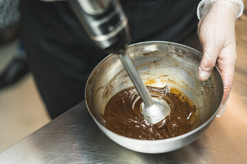 Top view of chocolate icing being blended in a metal bowl by a baker wearing transparent glove and black apron. . High quality photo