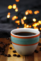 Colorful cup of black coffee with a cozy warm blanket on a wooden surface with the coffee beans and bokeh lights in the background.