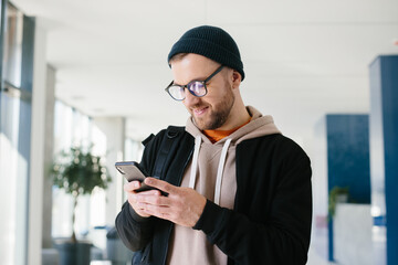 A young man in a casual outfit chats on a mobile phone while standing in the hall of an office building.