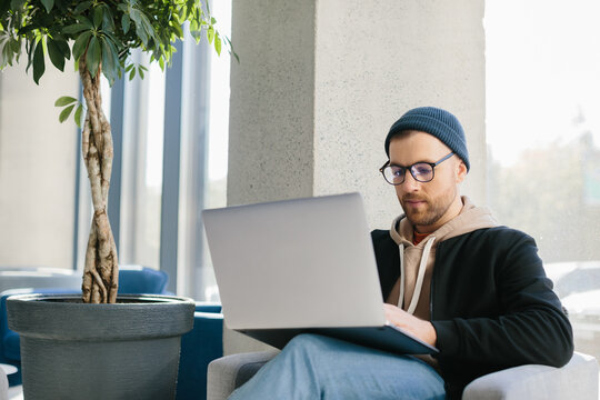 Young Handsome Freelancer Guy Working With Laptop In Office Space. A Man In Glasses And A Hat Is Holding A Laptop While Sitting In A Chair In The Hall.