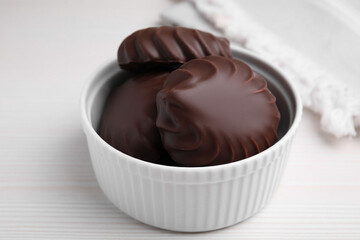 Delicious chocolate covered zephyrs in bowl on white wooden table, closeup