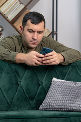 A 30-35-year-old man attentively looks through messages and videos in a smartphone, sitting behind a sofa, a serious expression on his face.