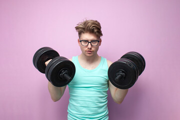 not athletic guy in glasses holds heavy dumbbells on a pink background and trains, motivated nerd goes in for sports