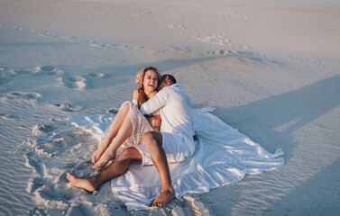 A bearded man kisses and hugs a laughing blonde woman sitting on white sand at sunset. A love story in the desert of newlyweds. Cheerful and loving people. Vacation and honeymoon.
