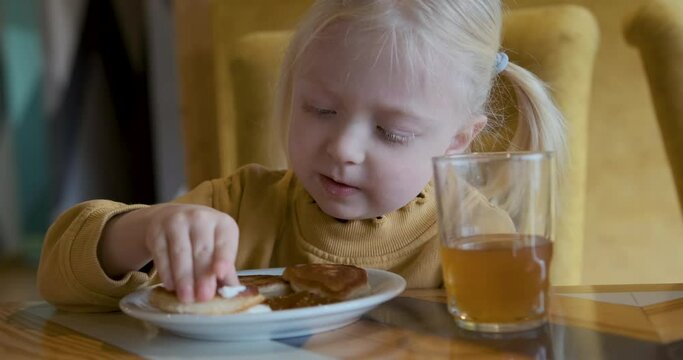 Close-up little fair-haired girl eating pancake with sour cream in cafe. Child licks cream from fritter and her fingers