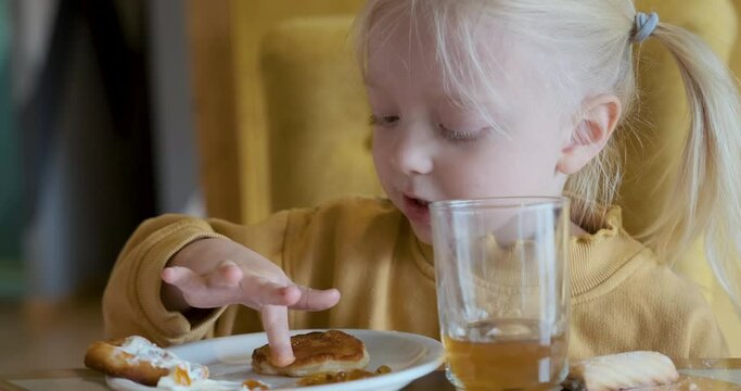 Little blonde girl enjoys eating pancakes with condensed milk licking fingers. Child in cafe with pastries
