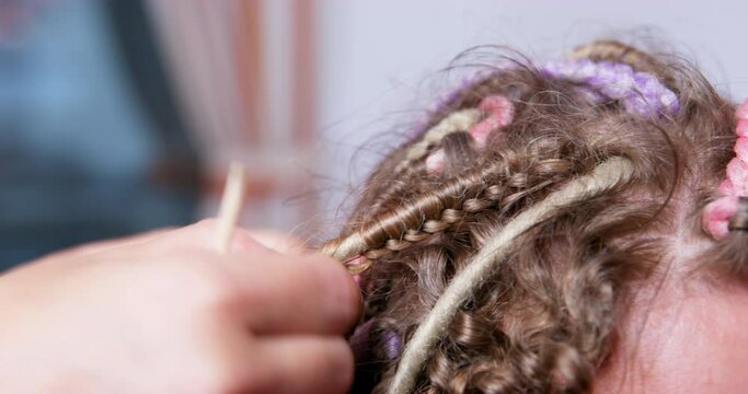 Hairdresser-stylist in a blue apron unwinds multicolored box braids to the client, using a special tool, side view close up. Professional master at work in a beauty salon.