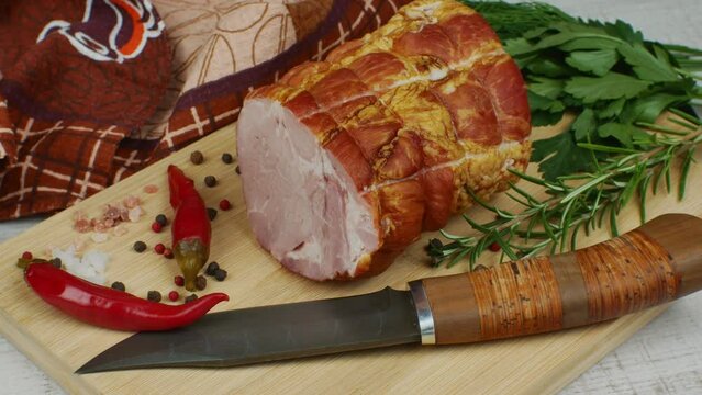 Traditional ham on a wooden cutting board with a carving knife, colorful allspice, red hot peppers, parsley, dill and basil The concept of delicious meat snacks from pork meat.