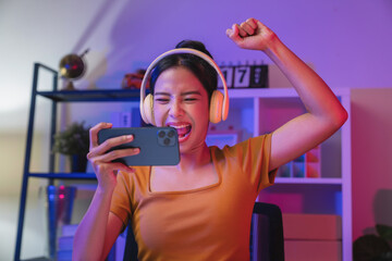 Excited Young Asian woman wearing headset and playing online game on smartphone at night modern house.