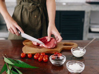 Woman with knife in hand cutting steak meat for frying in kitchen with salt pepper and other spices on table, red cherry tomatoes and herbs, dinner preparation.