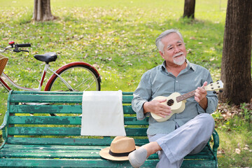 Happy smiling asian senior man with beard sitting on bench playing ukulele and singing a song in garden park outdoor. Musical and relaxation makes elder male happiness. Health care lifestyle concept.