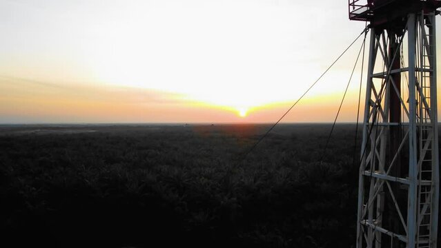 Cinematic 4K Drone Footage of Onshore Drilling Rig equipment structure for oil exploration and exploitation in the middle of jungle surrounded by palm oil trees during sunset and high oil price.