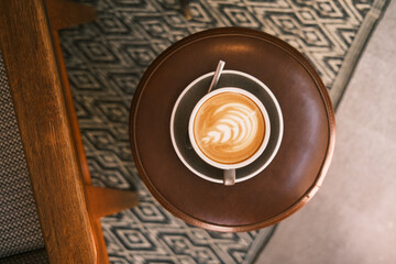 Café de barista con dibujo latte art hecho a mano y servido en taza y plato vintage sobre taburete de cuero y alfombra con patrón gométrico.
