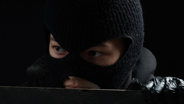 Masked female burglar in black gloves looks over a fence. Crime concept