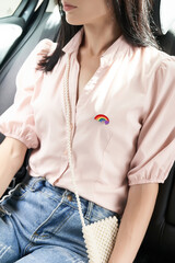 Cropped close-up shot of a young woman in a pink blouse and jeans with a rainbow pride flag lapel pin with a purple heart. A girl is sitting in a car with an LGBT flag pin. Front view.