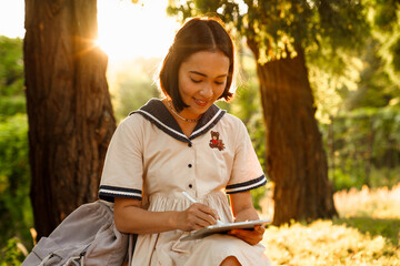 Young asian woman drawing on digital tablet with stylus while sitting in park