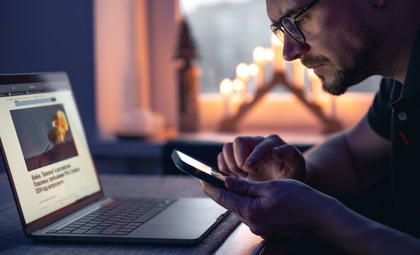 A man with a smartphone sits in front of a laptop late at night.