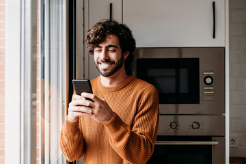 Happy young man using smart phone in kitchen at home