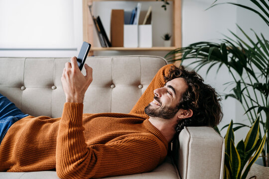 Happy young man using smart phone relaxing on sofa at home