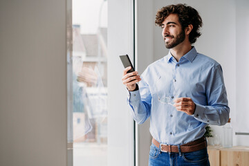 Happy businessman with smart phone looking through window in office