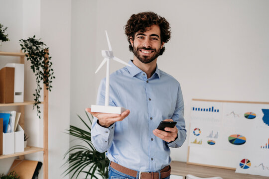 Happy engineer holding wind turbine and mobile phone in office - Powered by Adobe