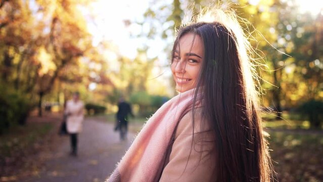 Rear view of girl with glass of coffee in hands go forward in park and waves her long beautiful dark hair to sides. Brunette joyfully skips and enjoy walk in autumn park with glass of coffee in hands.