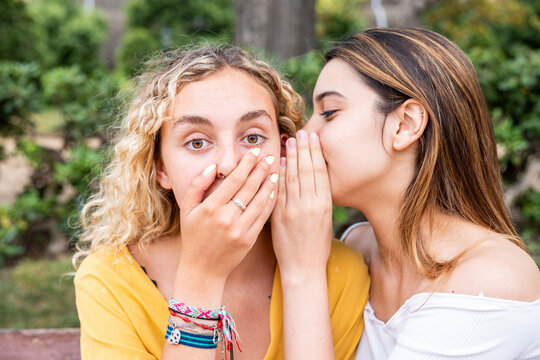 Woman whispering into shocked friend's ear in park