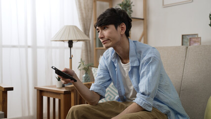 impatient asian Japanese man shaking head with dissatisfied look on face while channel surfing with...