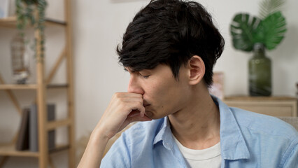 closeup view of a worried asian male propping face and gazing away into distance while pondering on a difficult decision in the living room at home